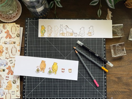 テーブル上に置かれたイラストが描かれた白い紙と文房具、動物のキャラクターが描かれたスケッチブックが見える