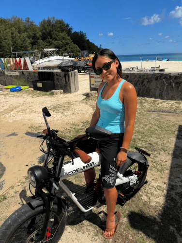 ビーチの近くでバイクに乗る女性。青いタンクトップと黒いパンツを着用し、サングラスをかけて晴れた日を楽しんでいる。背景には海と木々が見える。