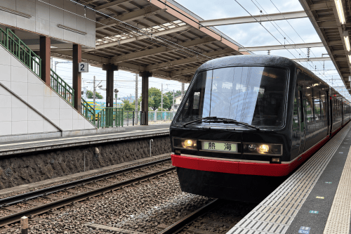 黒い車体の電車が駅に停車している様子。線路の近くにはプラットフォームがあり、周囲には緑のバリアフリーの階段が見える。