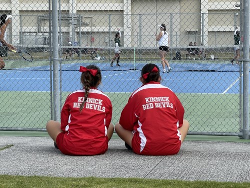 テニスコートの前で座っている2人の女性、赤いジャージに「Kinnick Red Devils」と書かれている