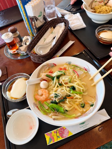 テーブルに置かれたラーメンと小皿料理の写真。ラーメンは海老、野菜、麺が入ったスープで、箸で食べる様子が見られる。