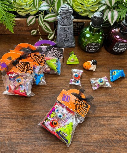 ハロウィンテーマのキャンディバッグが多数並んでいる画像。カラフルなキャンディ、小さなカボチャのオーナメントや目のモチーフが含まれている。背景には石の墓石やグリーンの植物がある。